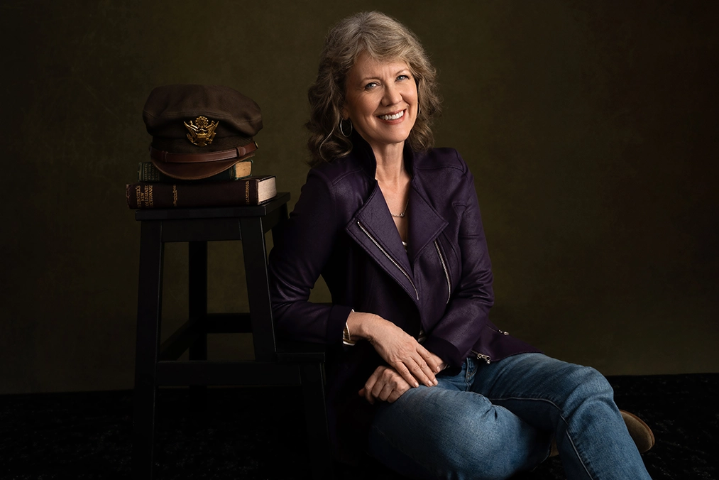 woman-in-purple-jacket-posing-next-to-military-cap-and-books-on-a-stool